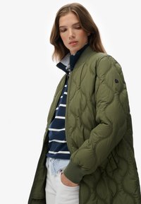 Giacca bomber trapuntata verde oliva con polsini a costine, chiusura con zip e tasche laterali, indossata sopra una camicia a righe blu navy e pantaloni bianchi.