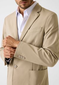 Blazer beige con texture liscia, design monopetto, rever a incavo e taschino sul petto. Presenta tre bottoni decorativi sui polsini.