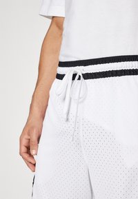 Hvide atletiske shorts med et tekstureret, perforeret stof. Har en elastisk talje med en sort snor og kontrasterende sorte detaljer.