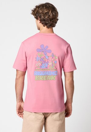 Homme portant un t-shirt rose avec un motif floral coloré et le texte "Blooming Break" au dos, associé à un pantalon beige.