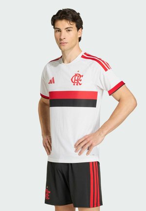 Young man models white soccer jersey with red and black stripes and black shorts with red stripes against plain background.