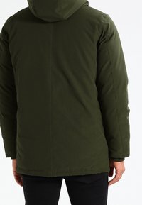 Dunkelgrüne Steppjacke mit Kapuze, die eine glatte Textur, gesteppte Nähte und einen geraden Schnitt aufweist, der bis zu den Hüften reicht.