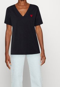 Czarna bawełniana koszulka T-shirt z dekoltem w serek, krótkimi rękawami, oraz haftowanym logo CK z czerwonym sercem na lewej piersi. Noszona z jasnymi dżinsami.