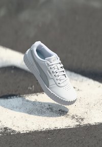 Białe buty sportowe z gładką skórzaną cholewką i teksturowaną gumową podeszwą, z subtelnymi szarymi akcentami i perforacjami zapewniającymi wentylację.