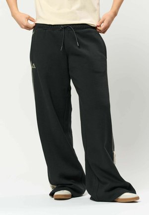 Person iført løse sorte sweatpants og hvide sneakers, der holder om taljen på sweatpants med begge hænder.