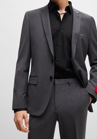 Giacca su misura grigia con bottoni neri, design a un petto, abbinata a una camicia nera con bottoni e colletto decorativo. Accenti rossi sulle maniche.