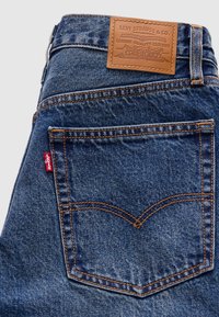 Πίσω τσέπη από μπλε τζιν denim με πορτοκαλί ραφές, κόκκινη ετικέτα Levi's και καφέ δερμάτινο ταμπελάκι Levi Strauss & Co. πάνω από τη μέση.