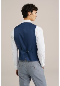 Chaleco azul marino con textura suave, con un ajustador de hebilla en la parte posterior, usado sobre una camisa blanca y pantalones ligeros de rayas grises.