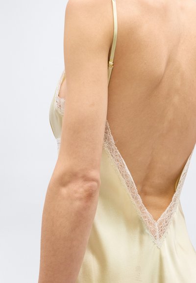 Πλάτη και χέρι γυναίκας που φοράει ανοιχτό κίτρινο σατέν φόρεμα με λεπτές τιράντες και δαντελωτή διακόσμηση κατά μήκος του βαθιού V-σχήματος ανοιχτής πλάτης.
