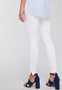 Femme portant un pantalon blanc ajusté avec de petites fentes à l'arrière et des sandales à talons hauts bleues, se tenant devant un fond uni.