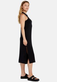 Vestito maxi nero senza maniche con spacco laterale, tessuto liscio, scollatura rotonda e vestibilità ampia, abbinato a sandali neri con fibbie.