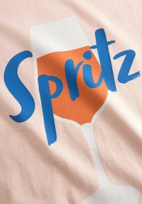 T-shirt in lichtroze stof met grote blauwe tekst "Spritz" en een illustratie van een wijnglas gevuld met oranje vloeistof.