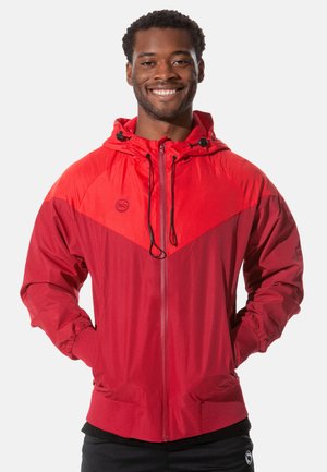 Red windbreaker jacket with a hood, featuring contrasting textured panels, elastic cuffs, and a zippered front. Logo on the chest.