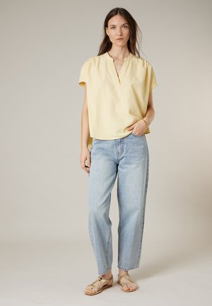 Woman wearing a yellow short-sleeve blouse, light blue jeans, and tan sandals, standing with one hand in her pocket on a plain background.