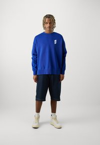 Felpa blu in cotone con texture a coste, dotata di una piccola patch con logo, abbinata a pantaloni corti in denim scuro e sneakers alte.