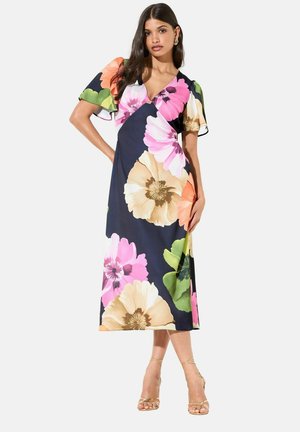 Žena v tmavě modrých zavinovacích midi šatech s velkými růžovými, béžovými a zelenými květinovými vzory a zlatými páskovými podpatky, stojící s jednou rukou na bok.