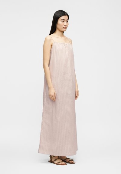 Mlada ženska z dolgimi črnimi lasmi, oblečena v brezrokavnik, dolgo obleko s roza belimi črtami in rjavimi ravnimi sandali.