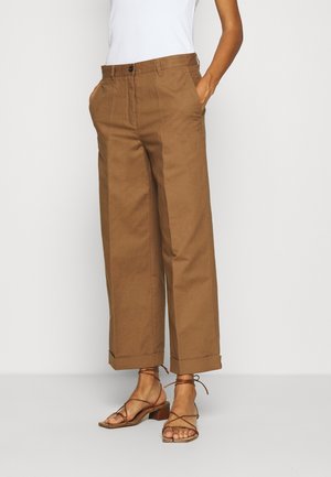 Vrouw draagt cropped, wijde bruine broek met omgeslagen zoom en bruine sandalen met hak en bandjes, handen in de zakken, staand tegen een effen achtergrond.