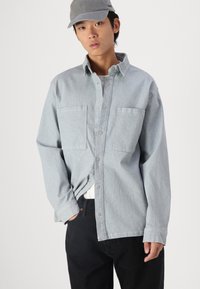 Lys blå, knappet skjorte med to brystlommer, laget av denim-lignende stoff. Modell iført svarte bukser og en grå caps. Enkel design.