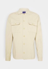 Camicia button-up beige in cotone con due tasche sul petto, chiusura con bottoni e colletto. Presenta bottoni gialli e maniche lunghe.