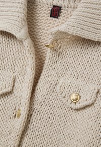 Cream-colored knitted cardigan with a textured pattern, gold hardware buttons, and a fold-over collar, featuring two front pockets.