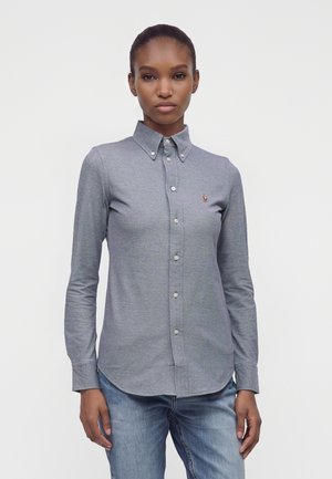 Femme portant une chemise grise cintrée à manches longues avec col et un jean bleu clair, debout devant un fond uni.