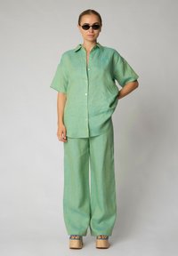 Camicia di lino verde menta con maniche corte e chiusura a bottoni, abbinata a pantaloni a gamba ampia dello stesso colore. Le scarpe con platform in legno completano l'outfit.