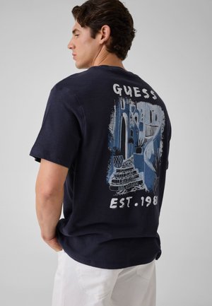 Muškarac nosi tamnoplavu Guess majicu s grafikom stiliziranih urbanih stepenica i zgrada i natpisom "GUESS EST. 1981" na leđima, u kombinaciji s bijelim hlačama.