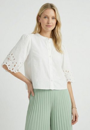 Vrouw draagt een witte blouse met knopen en oogjesmouwen en een lichtgroene geplooide rok, staand tegen een effen achtergrond.