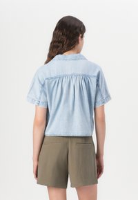 Ανοιχτό μπλε, κοντομάνικο τζιν πουκάμισο με  gather πίσω λεπτομέρεια, σε συνδυασμό με ελιές πράσινα σορτσάκια, βλέπεται από πίσω σε λευκό φόντο.