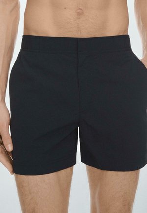 Pohled od pasu k polovinu stehen osoby ve stylových černých šortkách s elastickým pasem a bočními kapsami, stojící s uvolněnýma rukama.