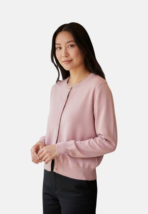 Donna con capelli lunghi e scuri che indossa un cardigan rosa chiaro con bottoni e pantaloni neri, in piedi davanti a uno sfondo chiaro e uniforme.