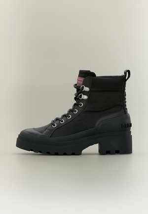 Botte noire montante à lacets avec semelle crantée épaisse, œillets métalliques, col rembourré et un patch bordeaux sur la languette.