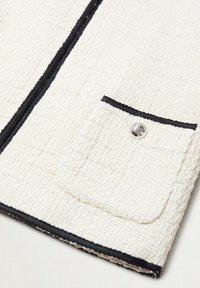 Tissu blanc texturé avec un léger éclat, présentant des coutures noires le long des bords et une petite poche avec un détail de bouton argenté.