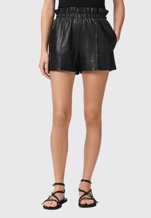 Kvinde iført sorte lædershorts med høj talje og sidelommer samt sorte flade sandaler med ankelstropper, stående mod en grå baggrund.