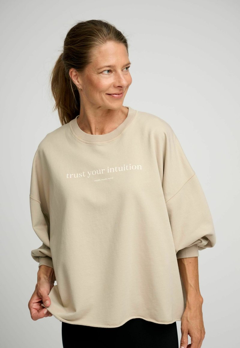 Beige oversized mikina s velkými rukávy, s nápisem "důvěřuj své intuici" v bílé barvě, vyrobená z texturované bavlněné směsi.