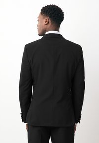 Black tailored suit jacket with a structured shoulder, single vent at the back, and buttoned cuffs. Smooth fabric with a crisp white shirt underneath.