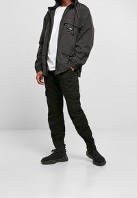 Black waterproof jacket with multiple pockets, paired with black cargo pants and black athletic shoes, displayed on a neutral background.