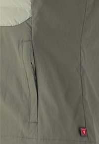 Olivgrüne Jacke mit glatter Textur, Reißverschluss-Tasche, kontrastierendem hellen Einsatz und rotem Markenlabel am Saum.