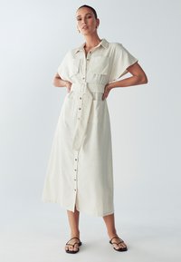 Beige Hemdblusenkleid mit kurzen Ärmeln, zwei Brusttaschen, tailliertem Gürtel und Midi-Länge, gefertigt aus einem strukturierten Stoff.