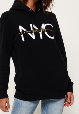 Personne portant un sweat à capuche noir avec « NYC » et un plus petit texte doré sur la poitrine, associé à un jean bleu clair, debout devant un fond uni.