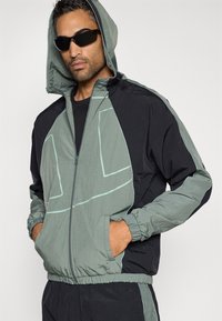 Kapucňová bunda v svetlozelenej a čiernej farbe, vyrobená z nylónu s hladkou textúrou. Obsahuje zips, elastické manžety a kontrastné geometrické vzory.