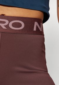 Burgunder røde treningsleggings med en bred midjebånd som har lyserosa "PRO"-merke. Materialet ser glatt og elastisk ut, designet for komfort.