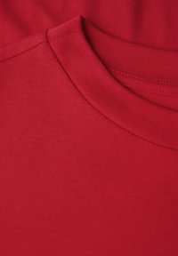 Röd bomullstyg med en slät textur. Har en rund halsringning och ett subtilt ribbat mönster längs kragen.