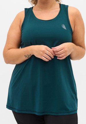 Frau mit lockigem Haar trägt ärmelloses, dunkel-türkisfarbenes Sporttanktop, die Hände vor dem Körper verschränkt, zeigt ein kleines Tattoo am linken Unterarm.