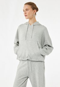 Jasnoszara bluza z kapturem wykonana z miękkiego, teksturowanego materiału. Posiada kaptur, przednią kieszeń oraz luźny krój z opuszczonymi szwami na ramionach.
