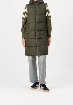 Personne portant une doudoune sans manches à capuche de couleur vert olive sur un pull à motifs, un jean bleu clair et des baskets noires à rayures blanches.