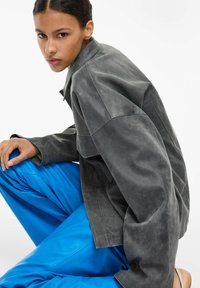 Hall nahktagi sileda lõdva lõikega, millel on nähtav õmbluse detail. Sobitatud säravsiniste, läikivate pükstega. Ühtlane sile tekstuur.