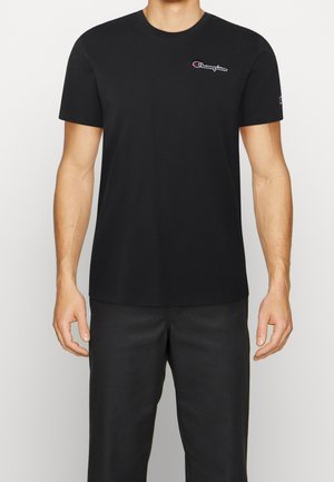 Man draagt een zwart kortemouws Champion T-shirt met logo op borst en mouw, gecombineerd met een zwarte broek, staand tegen een witte achtergrond.