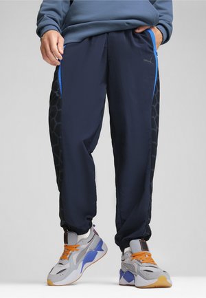 Puma trainingsbroek in marineblauw met een gestructureerd zwart hexagonaal patroon aan de zijkant, voorzien van blauwe accenten en elastische boorden. Draagt met chunky sneakers.